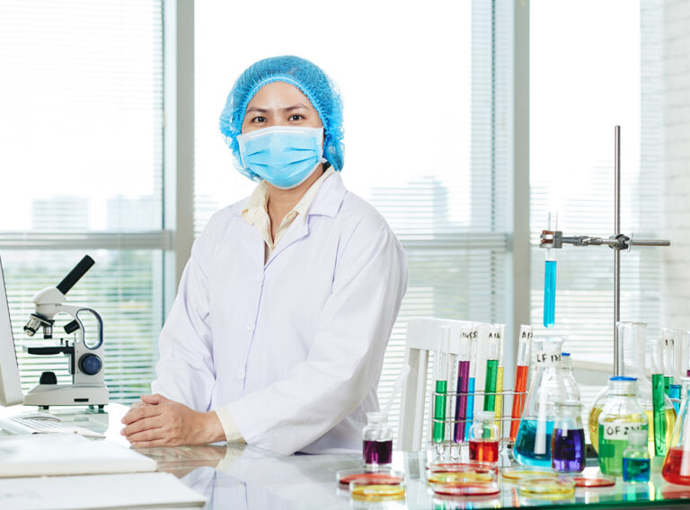 Portrait shot of confident Asian microbiologist wearing white coat and medical mask sitting at desk and posing for photography, interior of modern laboratory on background